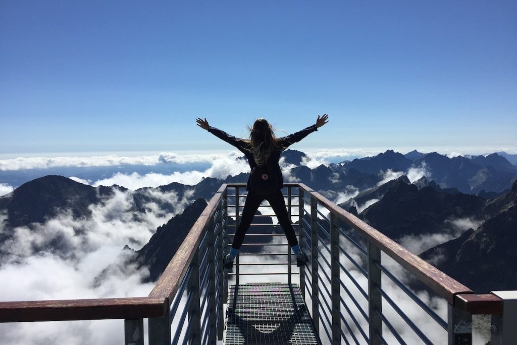 Person Standing on Hand Rails With Arms Wide Open Facing the Mountains and Clouds