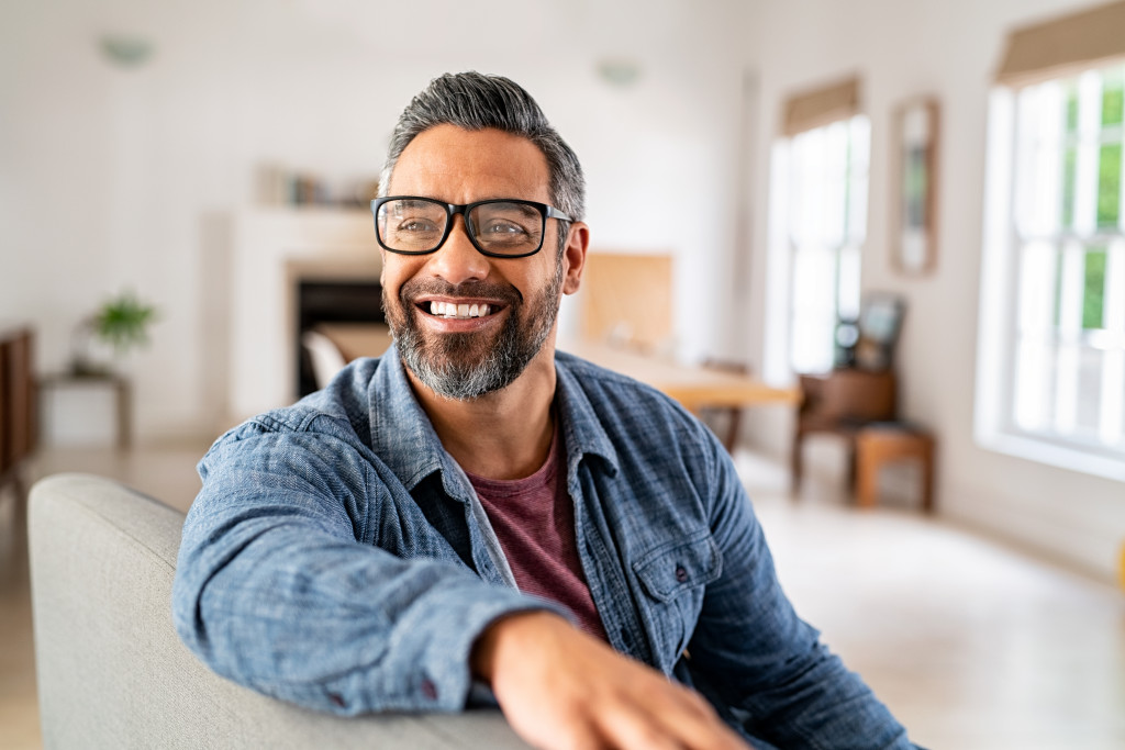 Mature man wearing eyeglasses smiling while sitting on a couch at home.